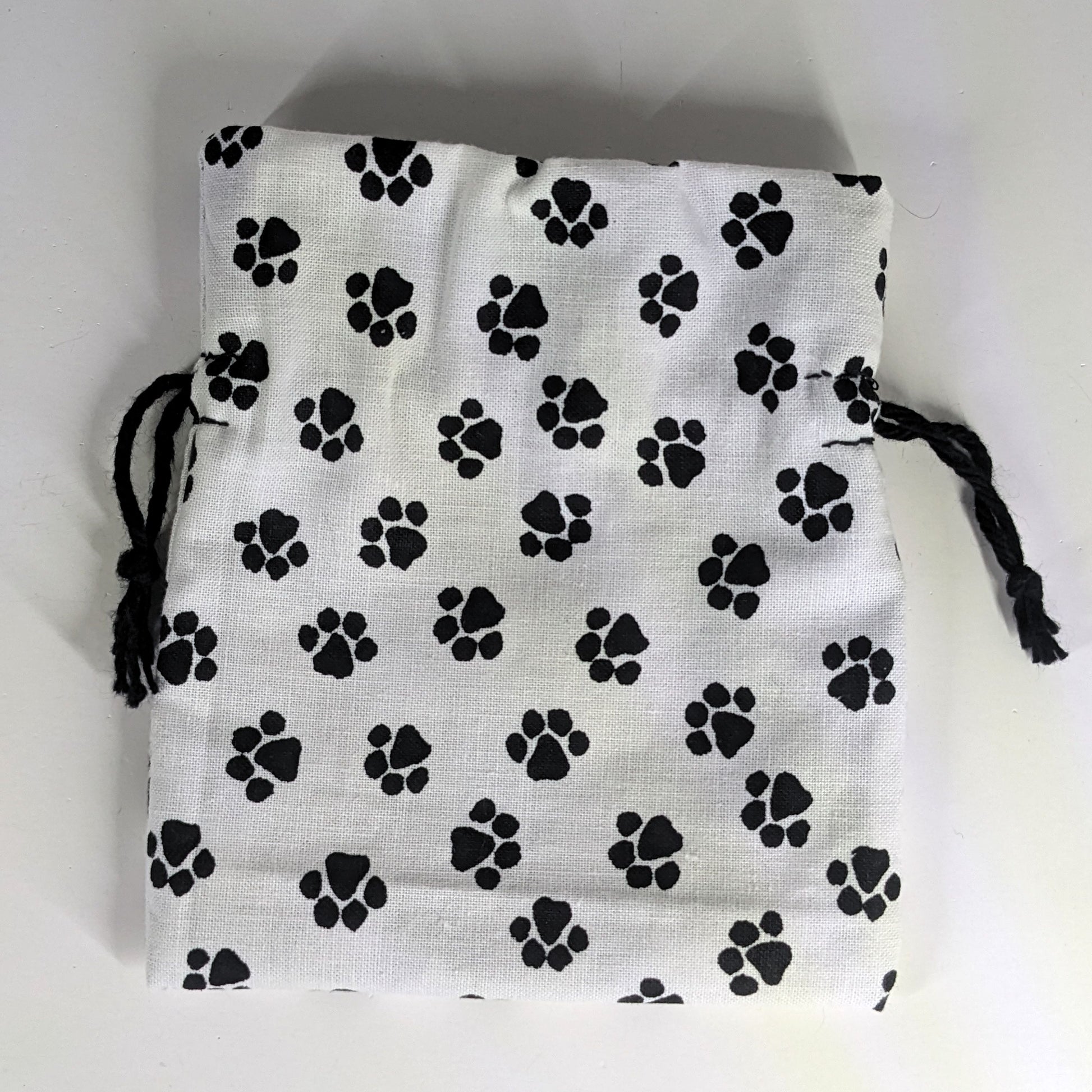 A small, handmade dice bag that is white with a black paw print lays flat on a white background.