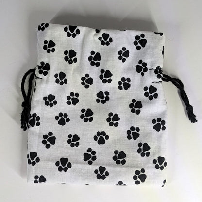 A small, handmade dice bag that is white with a black paw print lays flat on a white background.