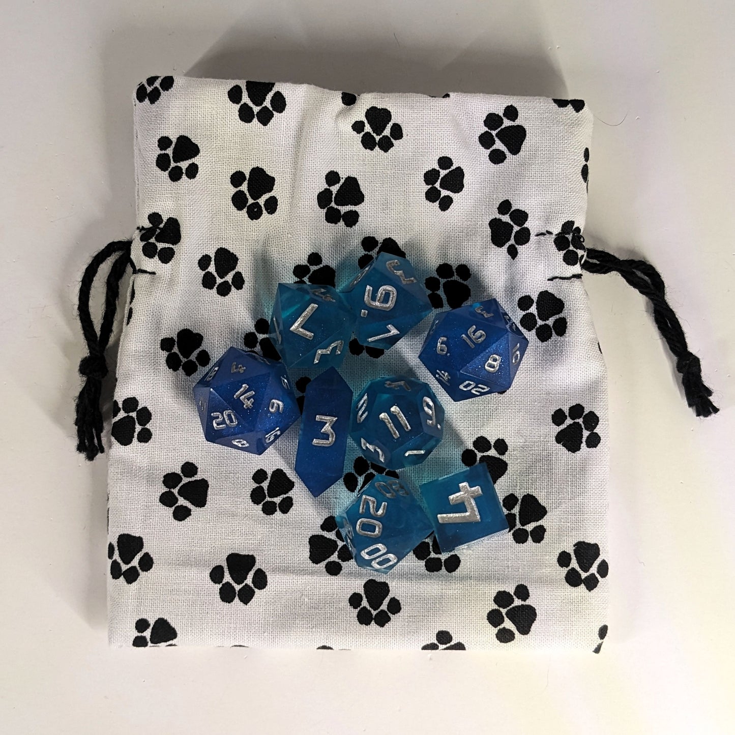 A small, handmade dice bag that is white with a black paw print pattern lays flat on a white background with a set of blue dnd dice sitting on top of the bag.