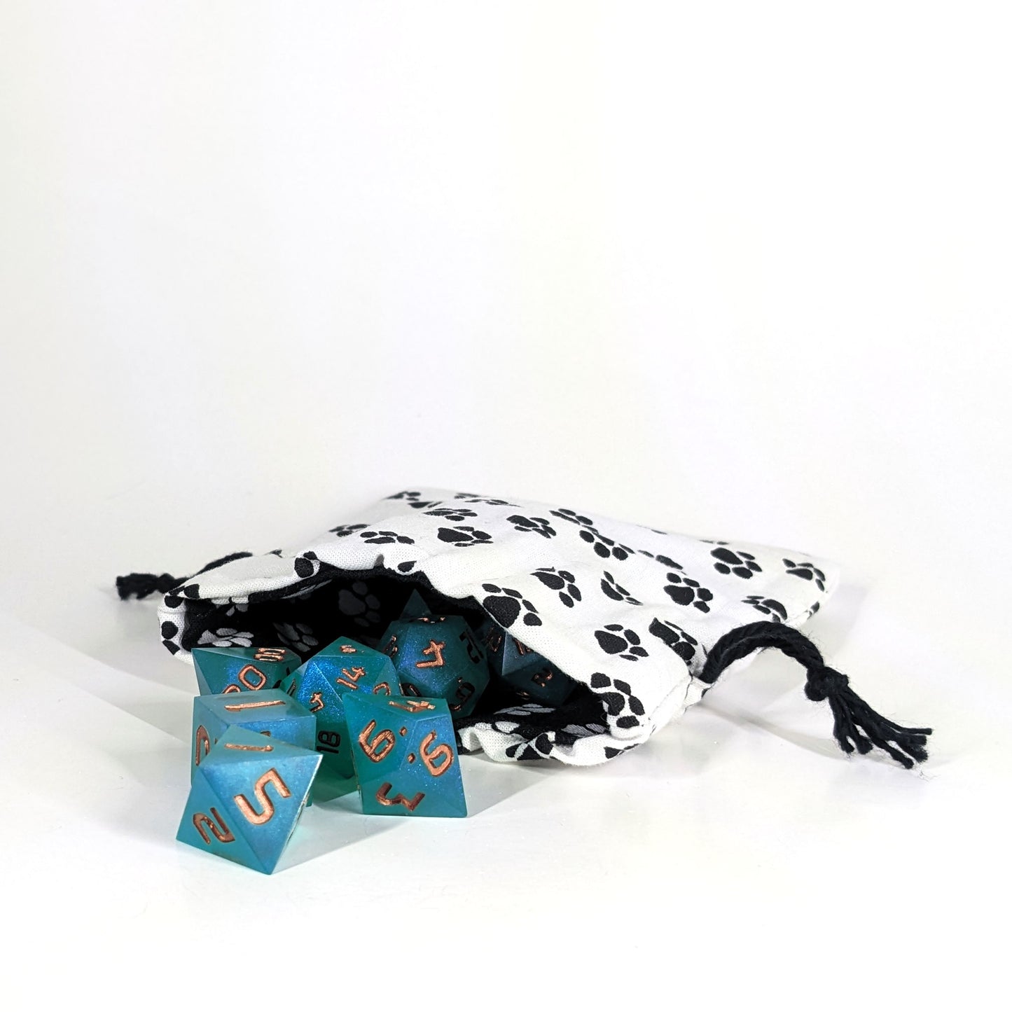 A small, handmade dice bag that is white with a black paw print pattern lays open showing a lining that is black with a white paw print pattern and teal dnd dice are spilling out of it on a white background.