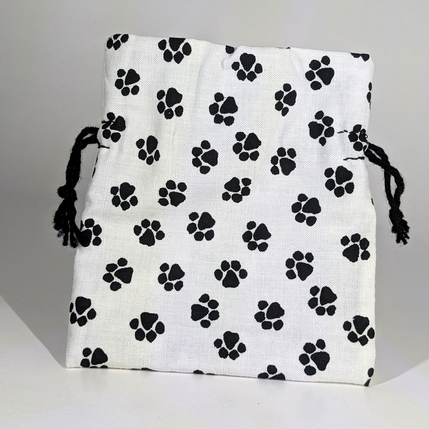 A small, handmade dice bag that is white with a black paw print pattern stands upright on a white background.