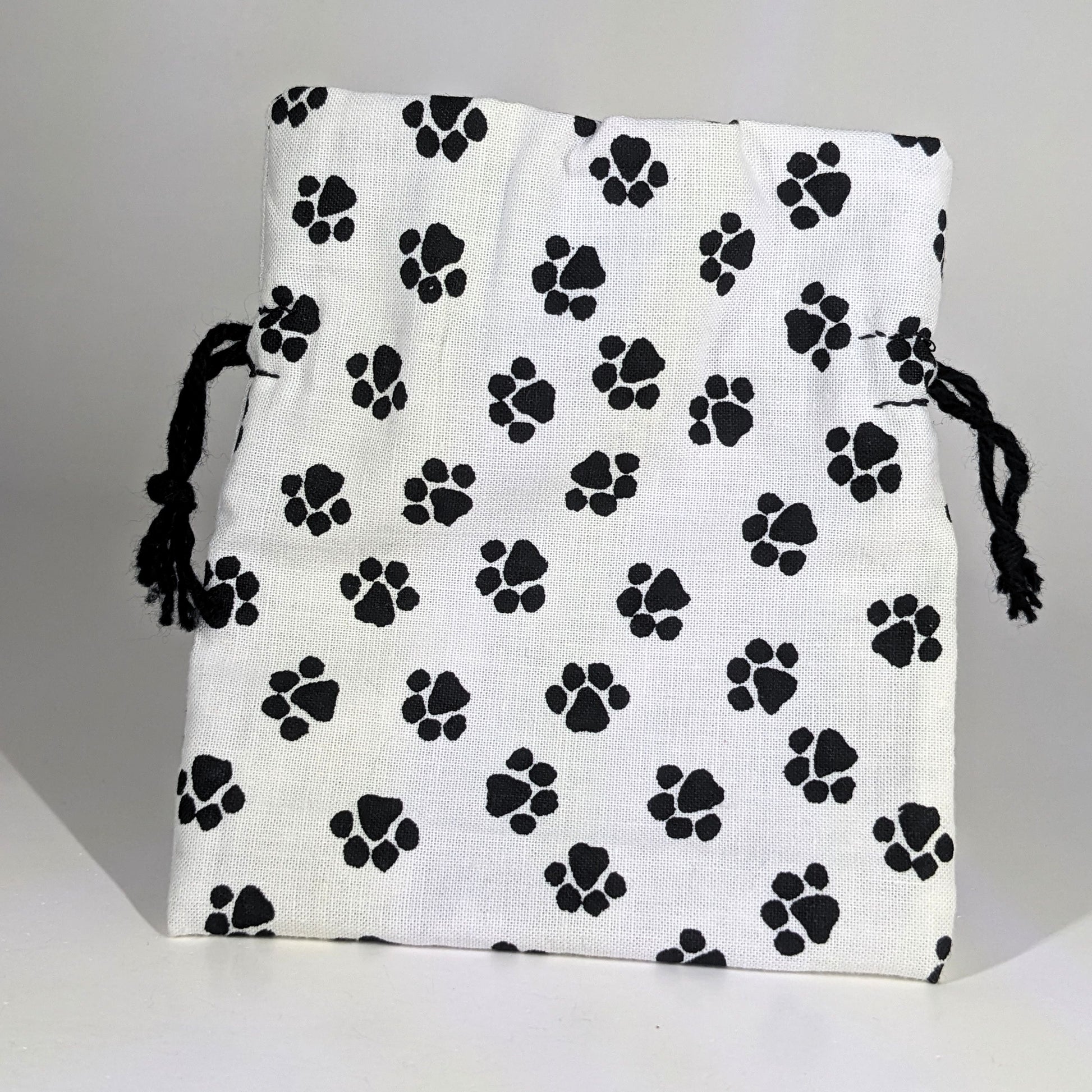 A small, handmade dice bag that is white with a black paw print pattern stands upright on a white background.