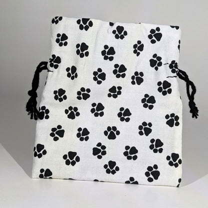 A small, handmade dice bag that is white with a black paw print pattern stands upright on a white background.