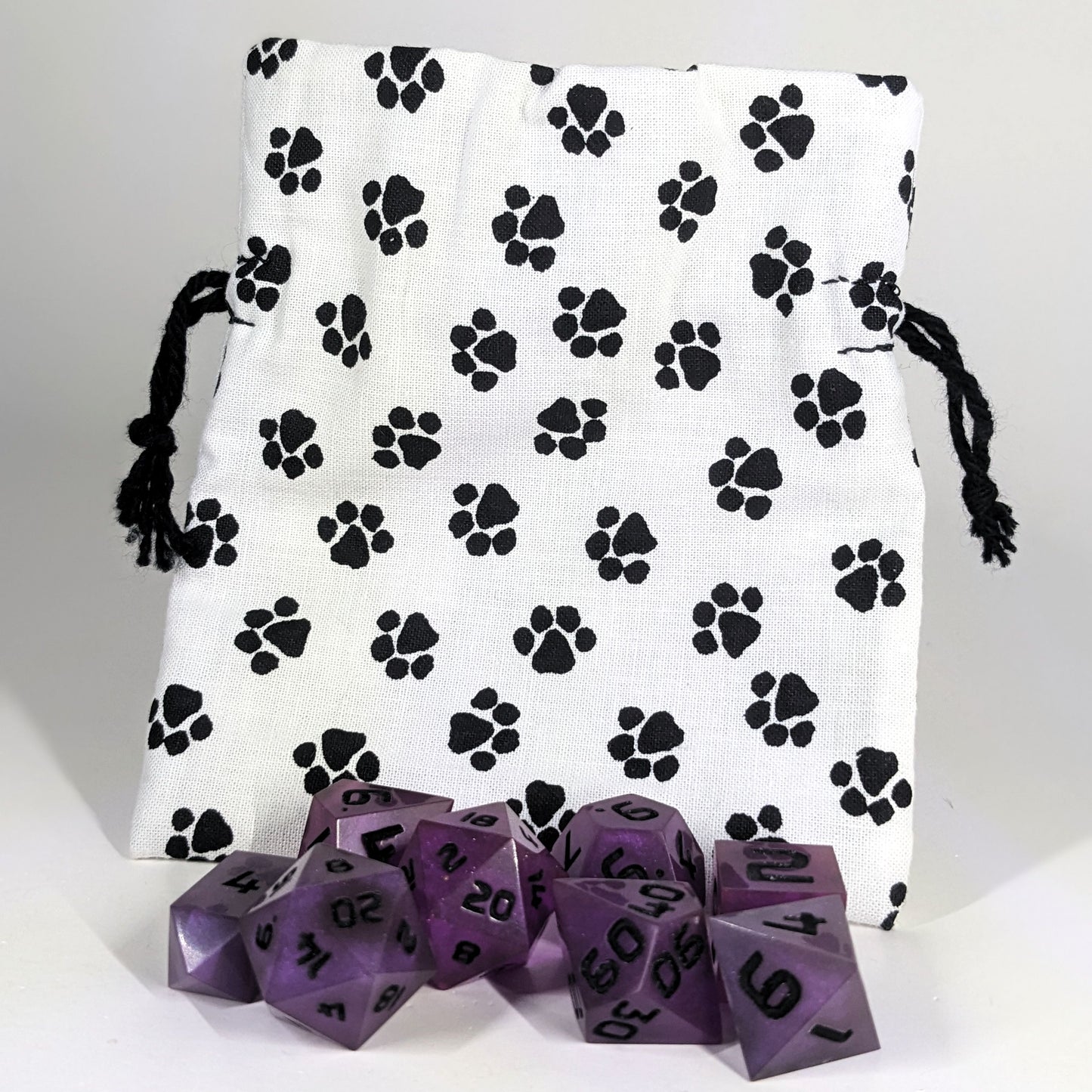 A small, handmade dice bag that is white with a black paw print pattern stands upright on a white background with a set of purple dnd dice sitting in front of the bag.