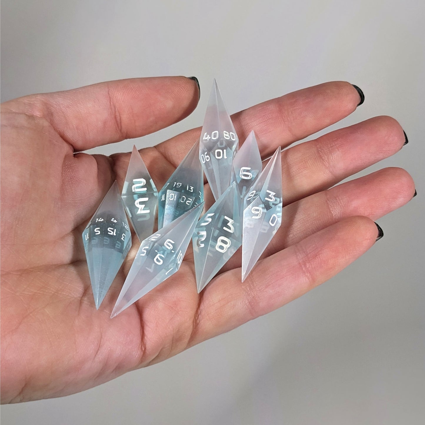 An 8 dice set of diamond style dice that are mixed blue and semitransparent white with opaque white numbers are held in a hand. The image shows that the set fits easily in a hand, making them overall smaller, though sharper, than a standard set of dnd dice.