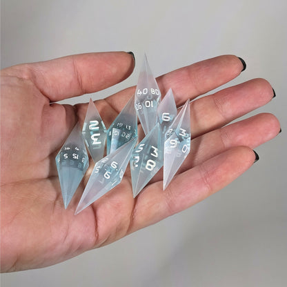 An 8 dice set of diamond style dice that are mixed blue and semitransparent white with opaque white numbers are held in a hand. The image shows that the set fits easily in a hand, making them overall smaller, though sharper, than a standard set of dnd dice.