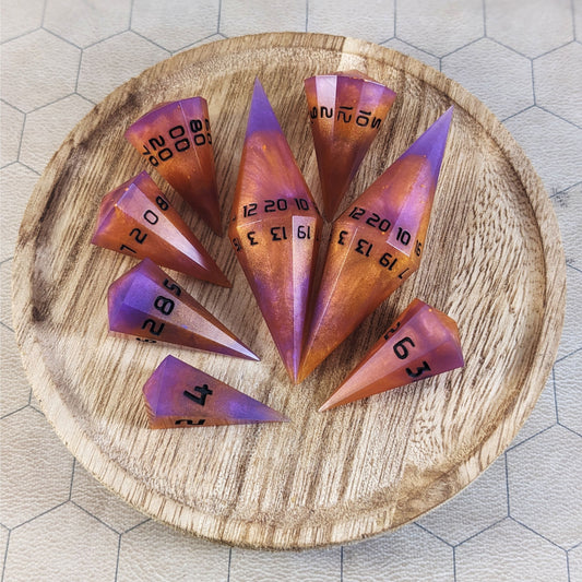 A complete set of dagger style resin dice that are purple and orange with black numbers are arranged on a wooden circle. The 2 d20s are next to each other with the d12 placed between them on top. The d6 is to the bottom right of the d20s and the rest of the dice are arranged along the left side, points angled toward the d20s.