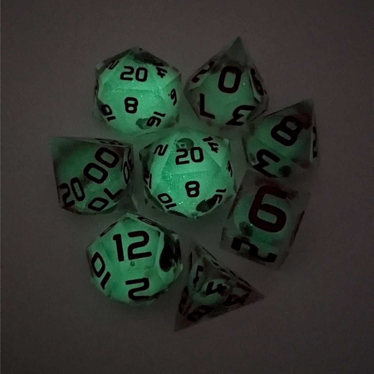 A complete set of 8 handmade artisan dice that have glow in the dark moon design inserts with red numbers. The DnD dice set is arranged in a circle and features 2 D20s, one of which is in the center surrounded by the other 7 dice on a white background. This photo was taken in the dark to show the spherical inserts glowing green in the dark.