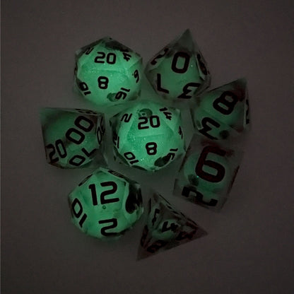 A complete set of 8 handmade artisan dice that have glow in the dark moon design inserts with red numbers. The DnD dice set is arranged in a circle and features 2 D20s, one of which is in the center surrounded by the other 7 dice on a white background. This photo was taken in the dark to show the spherical inserts glowing green in the dark.