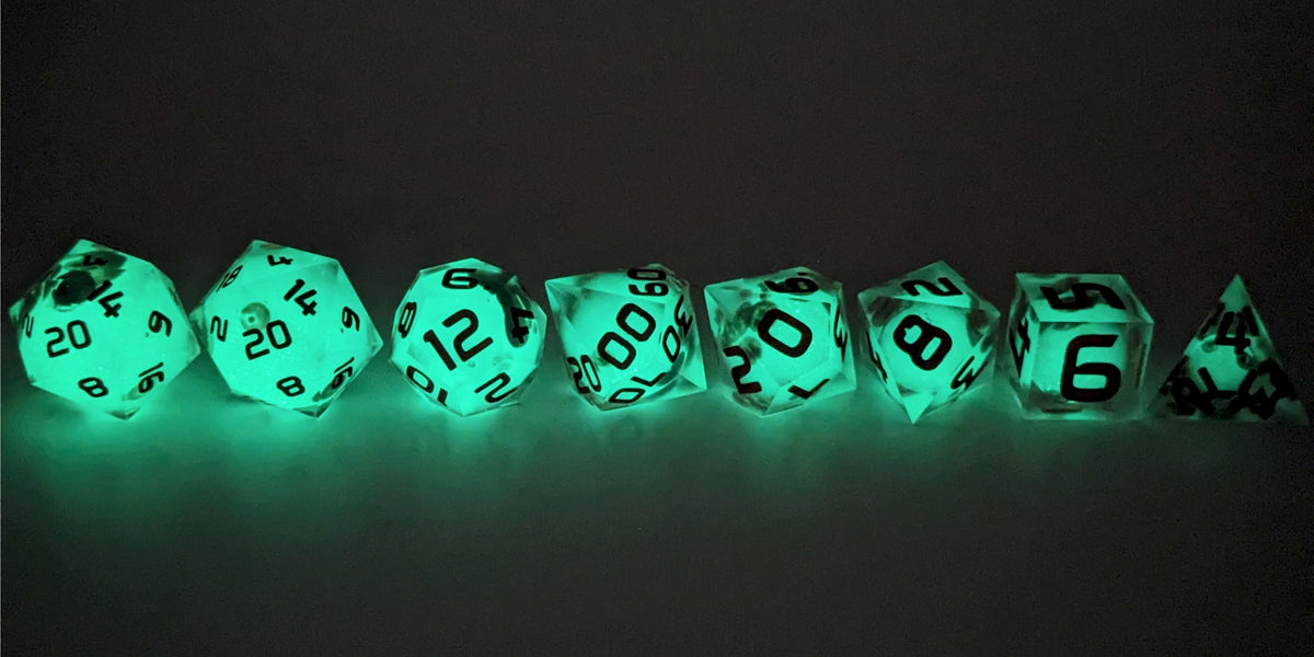 A straight on photo of a complete set of polyhedral dice that have glow in the dark moon design inserts with red numbers arranged in a straight line. They are lined up with the 2 d20s on the left going down to the d4 on the right side. The highest value number of each die is facing forward. This photo is taken in the dark to show the moon inserts glowing green in the dark.