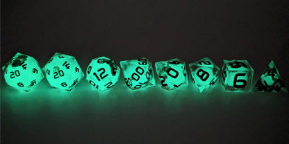 A straight on photo of a complete set of polyhedral dice that have glow in the dark moon design inserts with red numbers arranged in a straight line. They are lined up with the 2 d20s on the left going down to the d4 on the right side. The highest value number of each die is facing forward. This photo is taken in the dark to show the moon inserts glowing green in the dark.