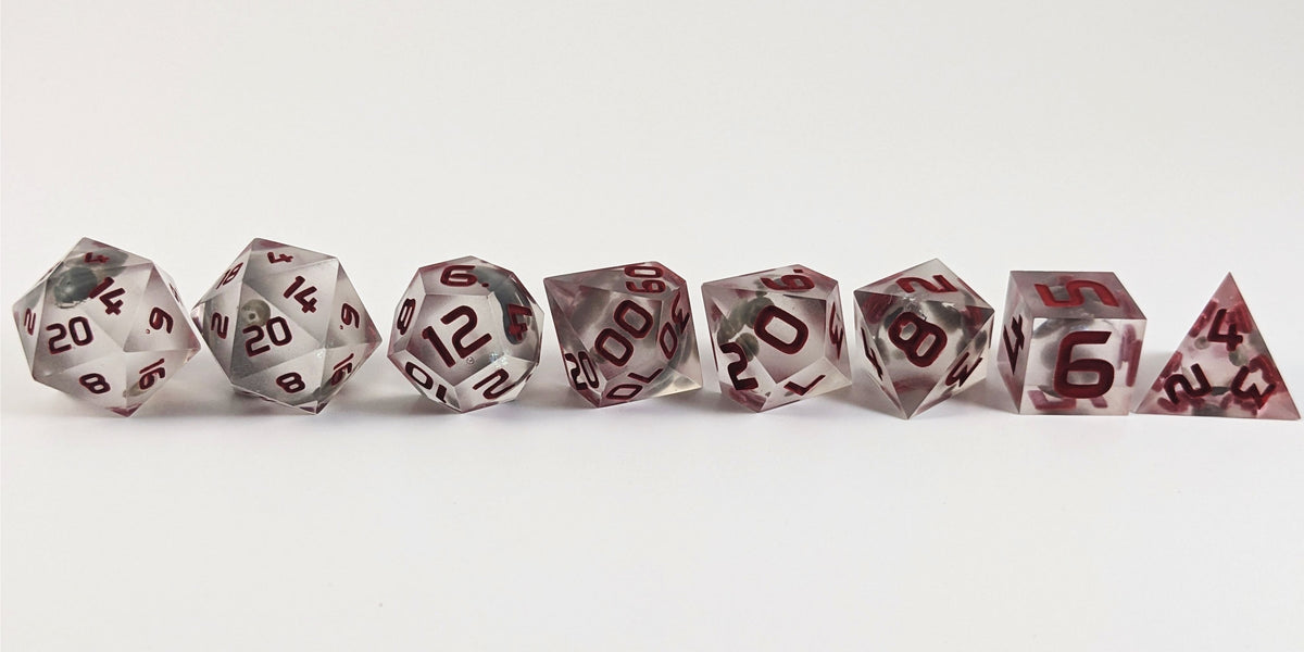 A straight on photo of a complete set of polyhedral dice that have glow in the dark moon design inserts with red numbers arranged in a straight line. They are lined up with the 2 d20s on the left going down to the d4 on the right side. The highest value number of each die is facing forward