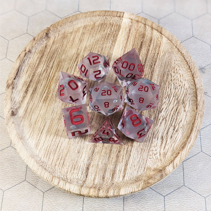 A complete set of 8 handmade artisan dice that have glow in the dark moon design inserts with red numbers. The DnD dice set is on a wooded circle and the dice are arranged in a circle and features 2 D20s, one of which is in the center surrounded by the other 7 dice.