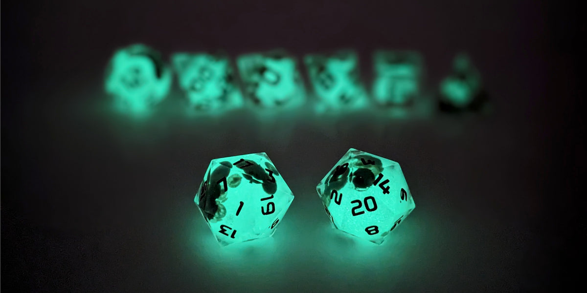 A straight on photo of a standard set of DnD dice that have glow in the dark moon design inserts with red numbers focusing on 2 d20s in the center. The d20 on the left has the 1 face centered and the d20 on the right has the 20 face centered. The other 6 dice are lined up in the background but are out of focus, while the d20s are in sharp focus. This photo is taken in the dark to show the moon spheres glowing green in the dark.