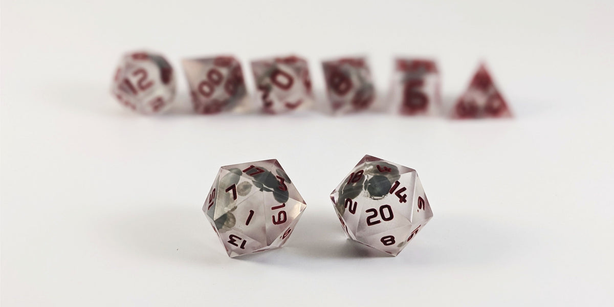 A straight on photo of a standard set of DnD dice that have glow in the dark moon design inserts with red numbers focusing on 2 d20s in the center. The d20 on the left has the 1 face centered and the d20 on the right has the 20 face centered. The other 6 dice are lined up in the background but are out of focus, while the d20s are in sharp focus.