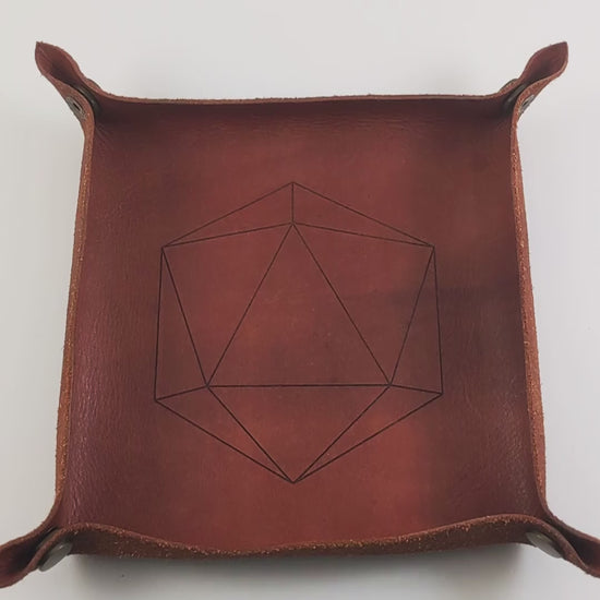 The video starts with a brown leather dice tray in view that has a d20 shape lasered into it. A hand appears from the bottom right corner holding a black and a white d12 each with gold numbers. The hand rolls the dice into the dice tray from the right.