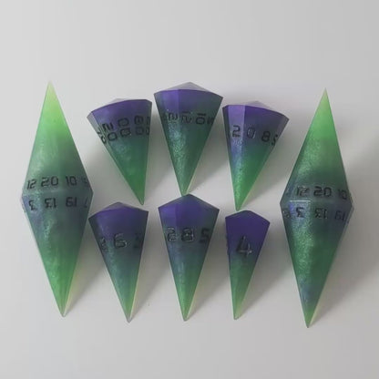 A video of a dagger style DnD dice set that is  green and purple with black numbers that glow in the dark is arranged with a d20 each on the right and left, with the d%, d12, and d10 along the top and the d6, d8, and d4 along the bottom, all in line with the d20s. The video shows the set transitioning from light to darkness to show the black numbers glowing green.