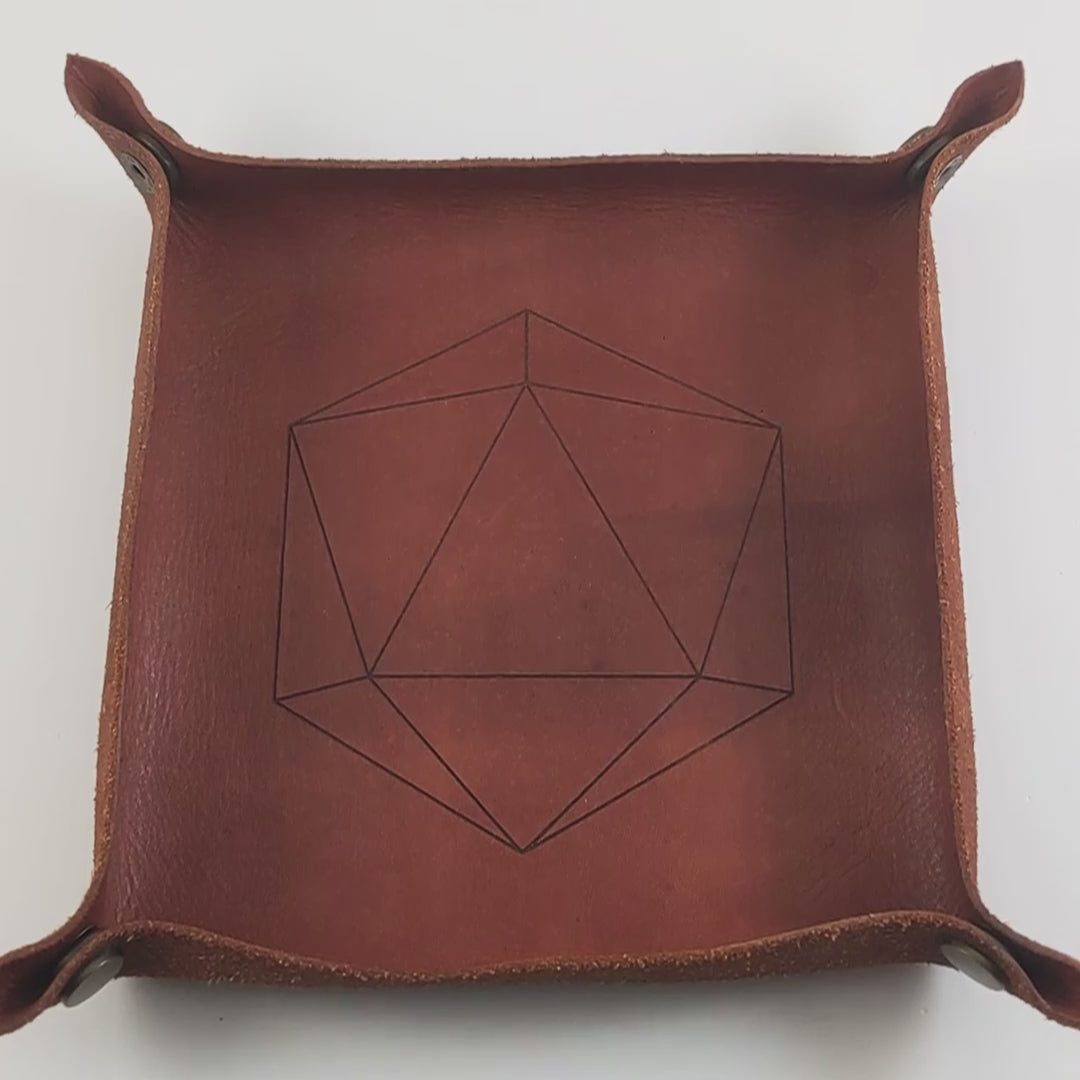 The video starts with a brown leather dice tray in view that has a d20 shape lasered into it. A hand appears from the bottom right corner holding a pair of yellow and teal d12s, one with white numbers and one with black numbers. The hand rolls the dice into the dice tray from the right.