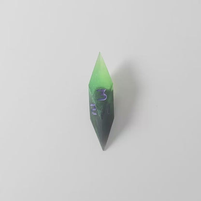 A video of a diamond d3 with swirls of black and green with purple numbers that glow in the dark on a white background. This transitions from light to darkness to show the purple numbers glowing green in the dark.