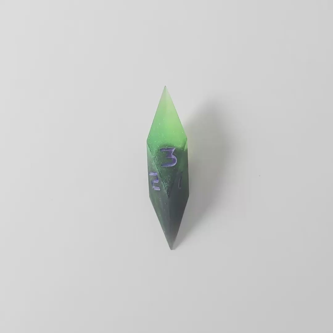 A video of a diamond d3 with swirls of black and green with purple numbers that glow in the dark on a white background. This transitions from light to darkness to show the purple numbers glowing green in the dark.