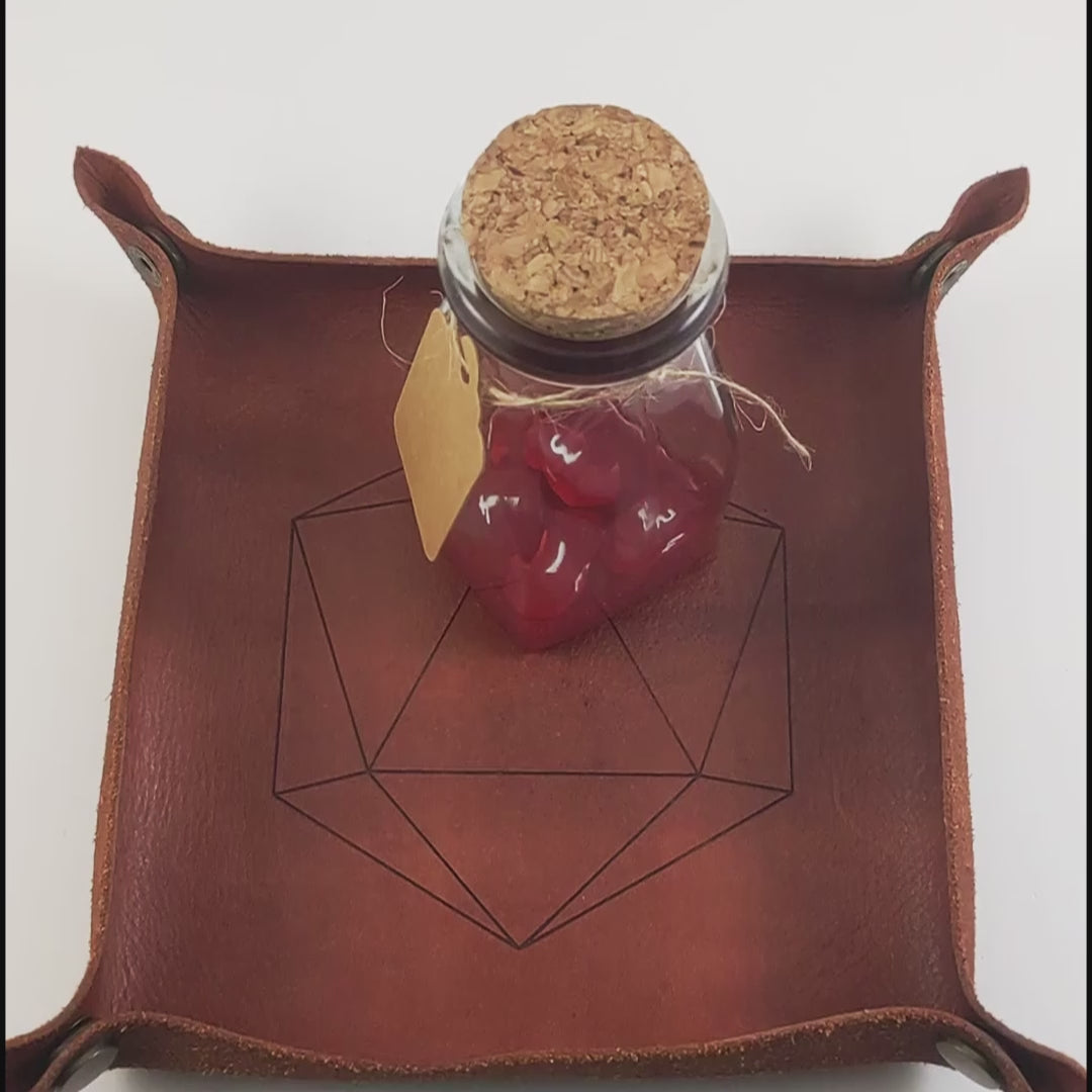 The video starts with a brown leather dice tray in view that has a d20 shape lasered into it and a potion bottle sitting on it. A hand appears from the right, holds the bottle up to the camera and flicks off the cork stopper with the index finger. The bottle is given a little shake and the four small red crystal dice are poured out of the bottle into the dice tray.