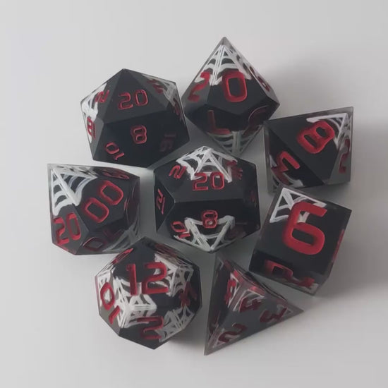 A video of a complete set of 8 handmade artisan dice that are black with hand painted white spider webs that glow in the dark and red numbers. The DnD dice set is arranged in a circle and features 2 D20s, one of which is in the center surrounded by the other 7 dice on a white background. This video goes from light to darkness to show the white webs glowing green.