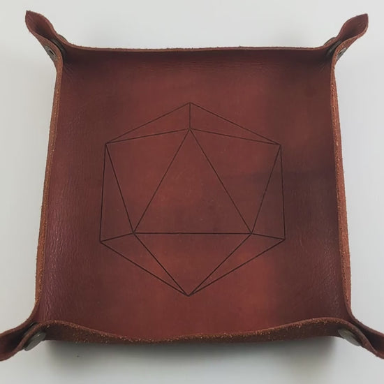 The video starts with a brown leather dice tray in view that has a d20 shape lasered into it. A hand appears from the bottom right corner holding a black d12 with white numbers and a white d12 with black numbers. The hand rolls the dice into the dice tray from the right.