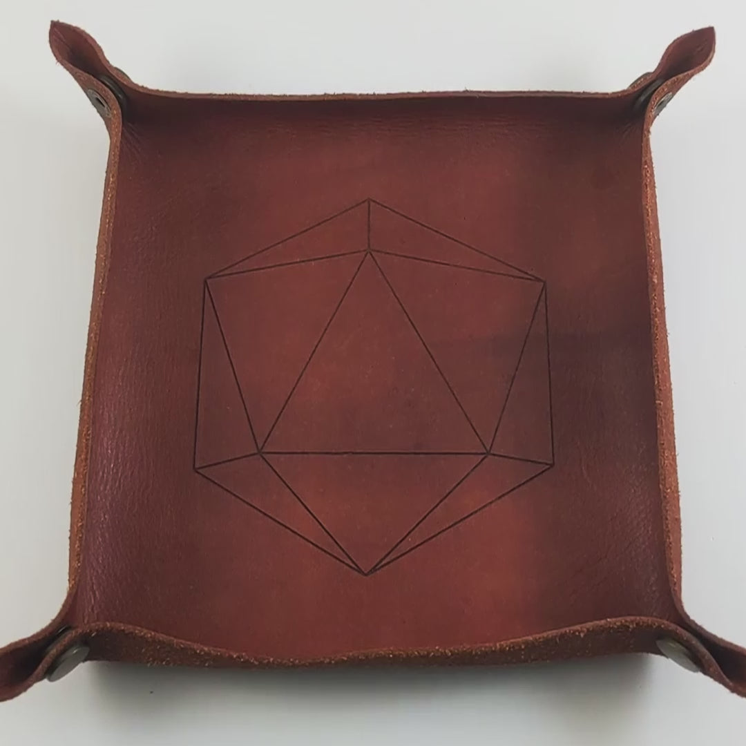 The video starts with a brown leather dice tray in view that has a d20 shape lasered into it. A hand appears from the bottom right corner holding a black d12 with white numbers and a white d12 with black numbers. The hand rolls the dice into the dice tray from the right.