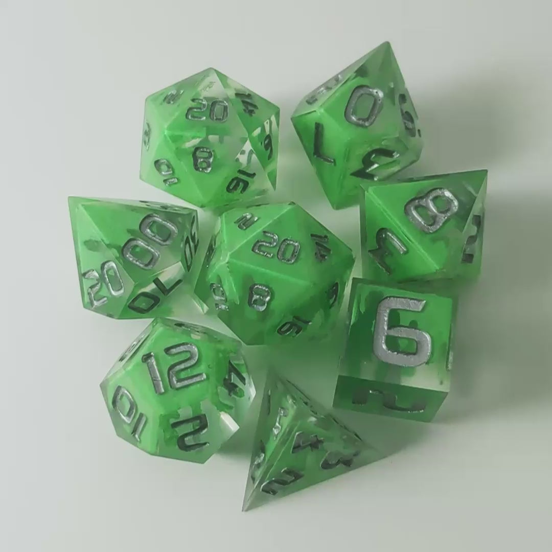 A video of a complete set of 8 handmade artisan dice that are clear with a hand painted green glow in the dark ooze design and silver numbers. The DnD dice set is arranged in a circle and features 2 D20s, one of which is in the center surrounded by the other 7 dice on a white background. This video transitions from light to darkness to show the ooze design glowing in the dark.