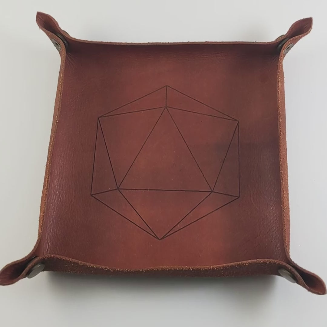 The video starts with a brown leather dice tray in view that has a d20 shape lasered into it. A hand appears from the bottom right corner holding a complete set of polyhedral dice that are gray with subtle marbling with painted gold lines along the edges a clear shell with ivory numbers. The hand rolls all of the dice into the tray from the right.