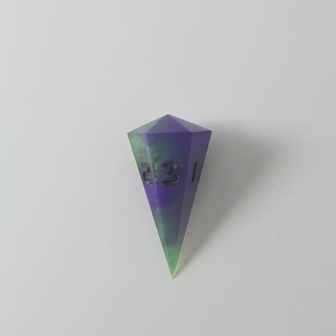 A video of a dagger d3 with swirls of purple and green with black numbers that glow in the dark on a white background. This transitions from light to darkness to show the black numbers glowing green in the dark.
