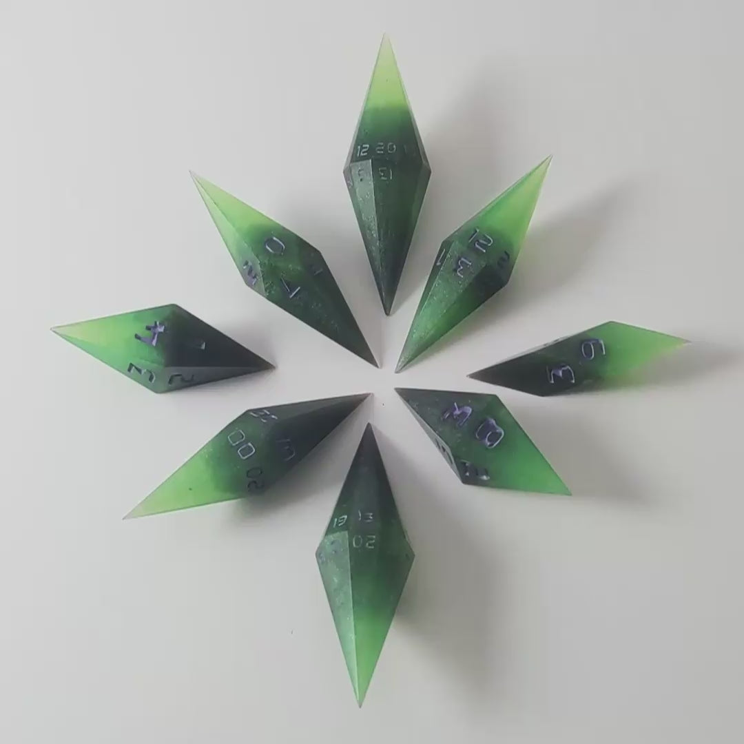A video of a complete set of diamond shaped resin dice featuring 2 d20s. These handmade dice are black and green with purple numbers that glow in the dark and are clustered in a circle with the bottom points angled toward a central point on a white background. This video transitions from being in the light to the dark to show the purple numbers glowing green in the dark.