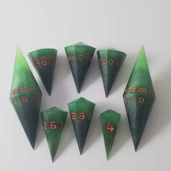 A video of a dagger style DnD dice set that is  green and black with orange numbers that glow in the dark is arranged with a d20 each on the right and left, with the d%, d12, and d10 along the top and the d6, d8, and d4 along the bottom, all in line with the d20s. The video shows the set transitioning from light to darkness to show the orange numbers glowing green.