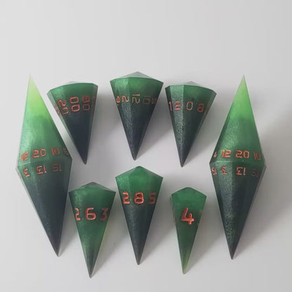 A video of a dagger style DnD dice set that is  green and black with orange numbers that glow in the dark is arranged with a d20 each on the right and left, with the d%, d12, and d10 along the top and the d6, d8, and d4 along the bottom, all in line with the d20s. The video shows the set transitioning from light to darkness to show the orange numbers glowing green.