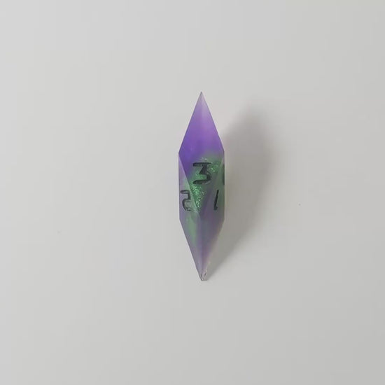 A video of a diamond d3 with swirls of purple and green with black numbers that glow in the dark on a white background. This transitions from light to darkness to show the black numbers glowing green in the dark.