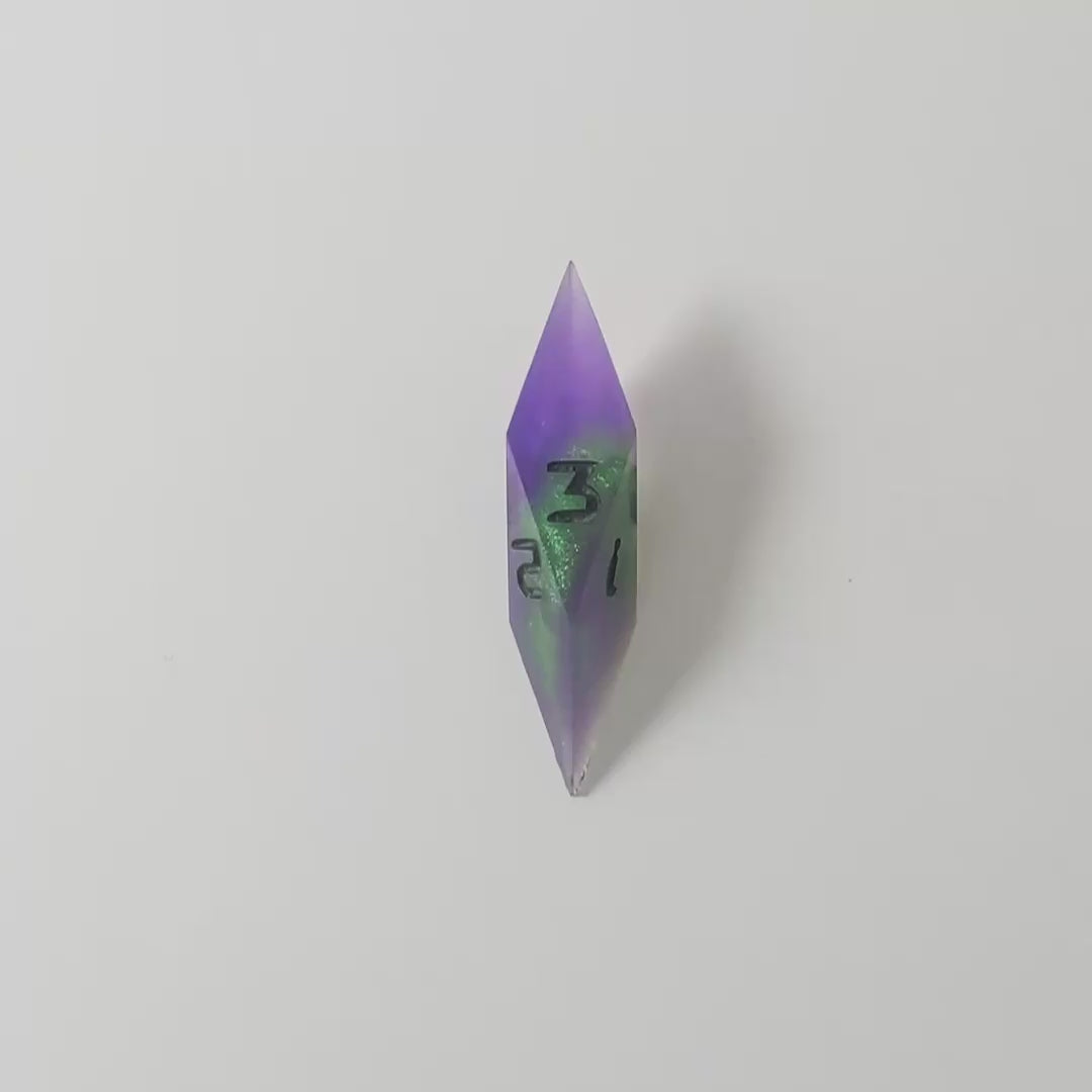 A video of a diamond d3 with swirls of purple and green with black numbers that glow in the dark on a white background. This transitions from light to darkness to show the black numbers glowing green in the dark.