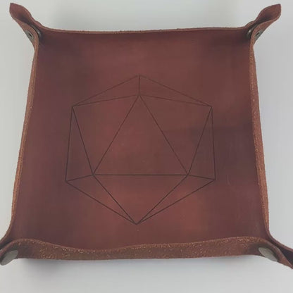 The video starts with a brown leather dice tray in view that has a d20 shape lasered into it. A hand appears from the bottom right corner holding a complete set of polyhedral dice that are black with hand painted white spider webs that glow in the dark with red numbers. The hand rolls all of the dice into the tray from the right.