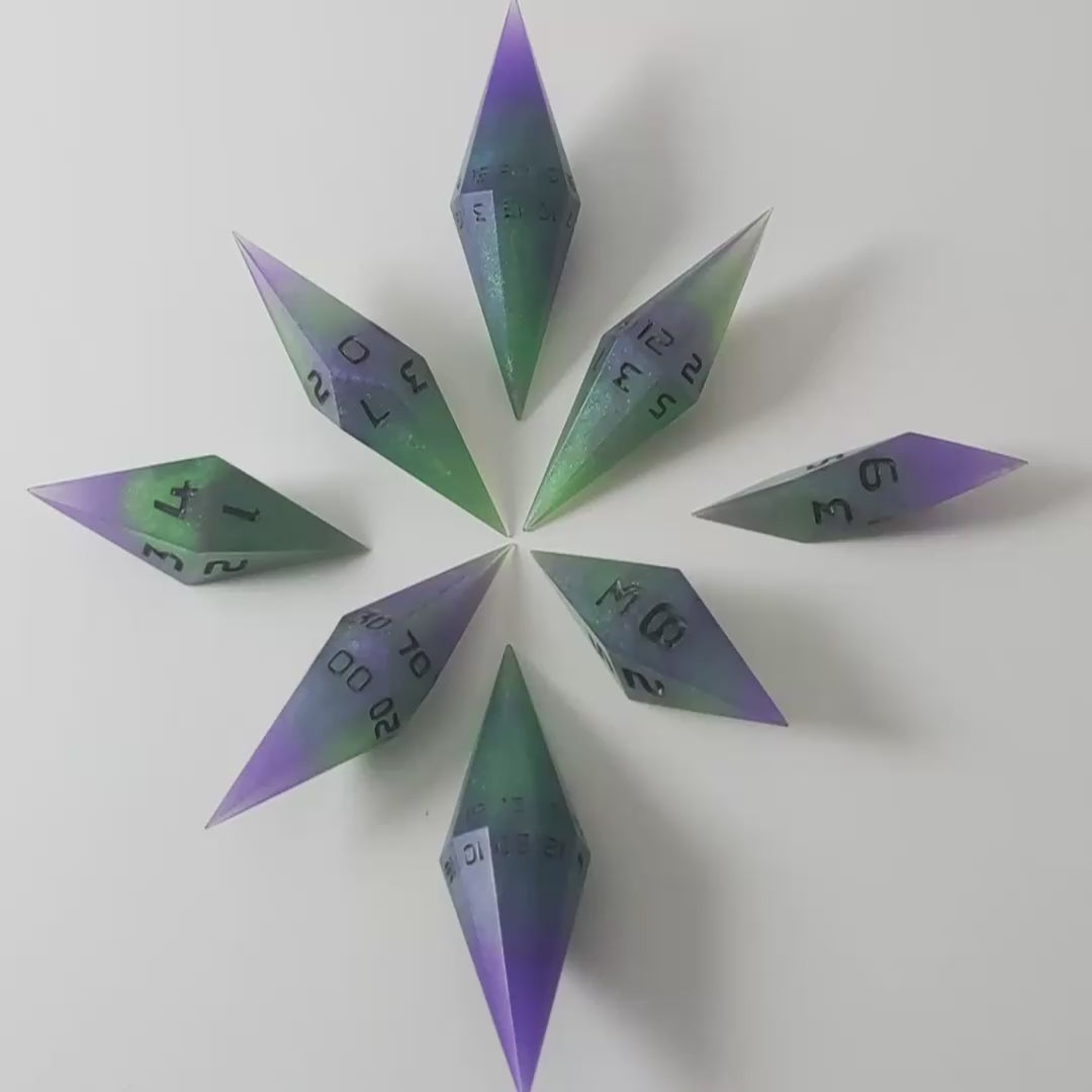 A video of a complete set of diamond shaped resin dice featuring 2 d20s. These handmade dice are purple and green with black numbers that glow in the dark and are clustered in a circle with the bottom points angled toward a central point on a white background. This video transitions from being in the light to the dark to show the black numbers glowing green in the dark.