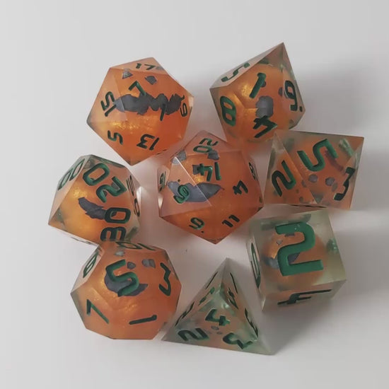 A video of a complete set of 8 handmade artisan dice that have jack-o-lantern inserts whose faces glow in the dark with dark green numbers. The DnD dice set is arranged in a circle and features 2 D20s, one of which is in the center surrounded by the other 7 dice on a white background. The dice are arranged so the jack-o-lantern faces are facing as upwards as possible. The video transitions from light to darkness to show the faces glowing green in the dark.