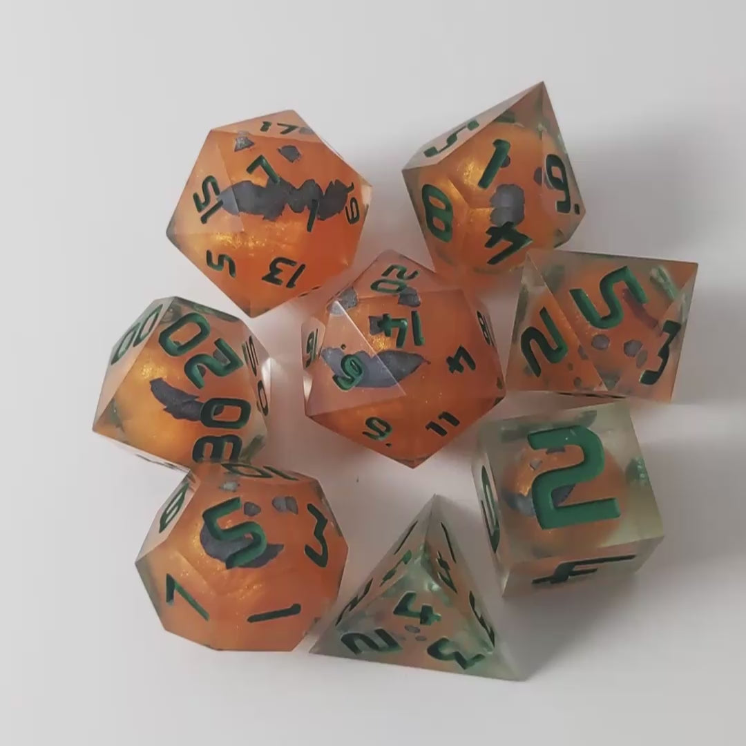 A video of a complete set of 8 handmade artisan dice that have jack-o-lantern inserts whose faces glow in the dark with dark green numbers. The DnD dice set is arranged in a circle and features 2 D20s, one of which is in the center surrounded by the other 7 dice on a white background. The dice are arranged so the jack-o-lantern faces are facing as upwards as possible. The video transitions from light to darkness to show the faces glowing green in the dark.