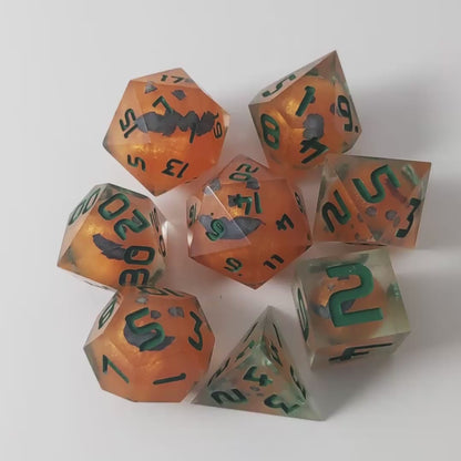 A video of a complete set of 8 handmade artisan dice that have jack-o-lantern inserts whose faces glow in the dark with dark green numbers. The DnD dice set is arranged in a circle and features 2 D20s, one of which is in the center surrounded by the other 7 dice on a white background. The dice are arranged so the jack-o-lantern faces are facing as upwards as possible. The video transitions from light to darkness to show the faces glowing green in the dark.