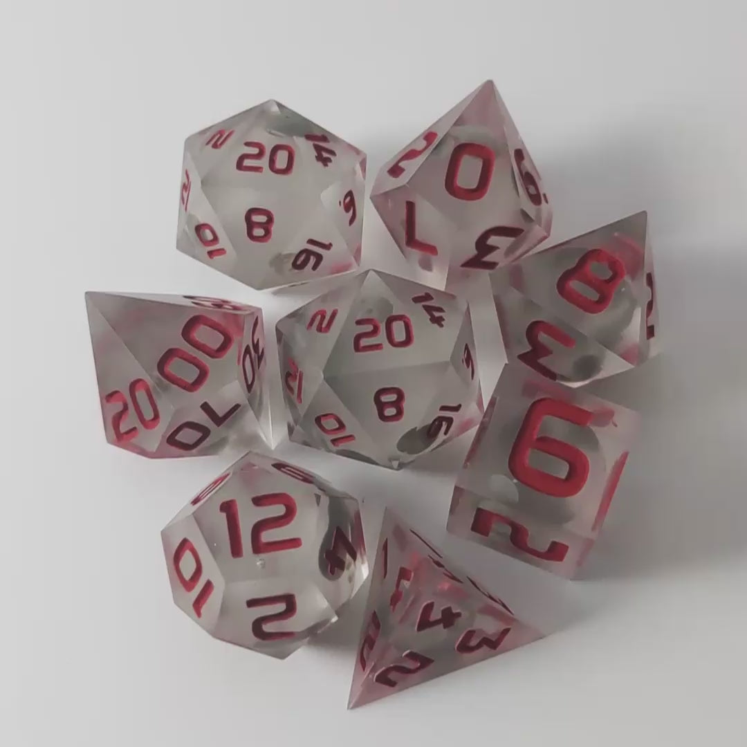 A video of a complete set of 8 handmade artisan dice that have glow in the dark moon design inserts with red numbers. The DnD dice set is arranged in a circle and features 2 D20s, one of which is in the center surrounded by the other 7 dice on a white background. This video was transitions from light to darkness to show the spherical inserts glowing green in the dark.