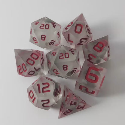 A video of a complete set of 8 handmade artisan dice that have glow in the dark moon design inserts with red numbers. The DnD dice set is arranged in a circle and features 2 D20s, one of which is in the center surrounded by the other 7 dice on a white background. This video was transitions from light to darkness to show the spherical inserts glowing green in the dark.