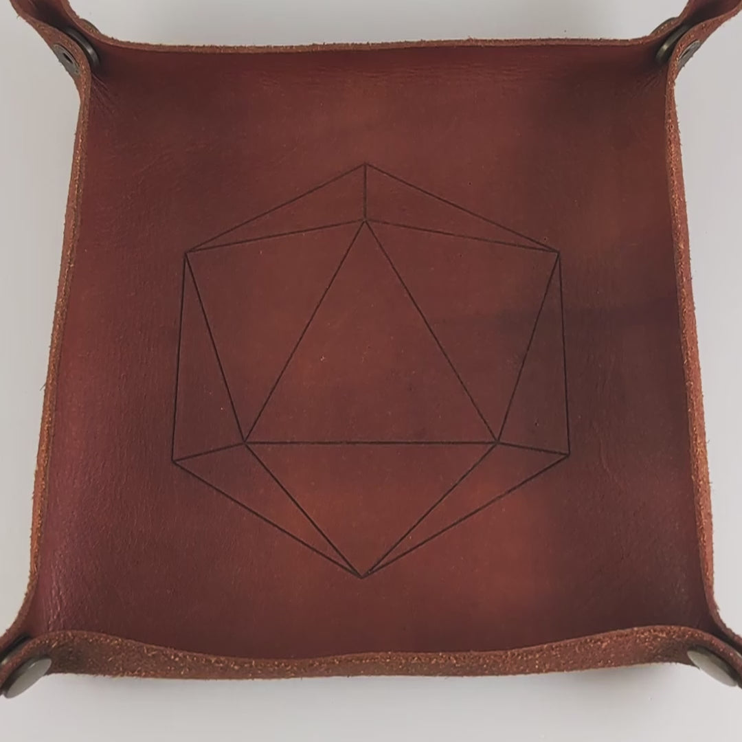 The video starts with a brown leather dice tray in view that has a d20 shape lasered into it. A hand appears from the bottom right corner holding a complete set of dagger dice that are maroon, gold, and dark silver layers with white numbers. The hand rolls all of the dice into the tray from the right. One of the d20s rolls a natural 20.